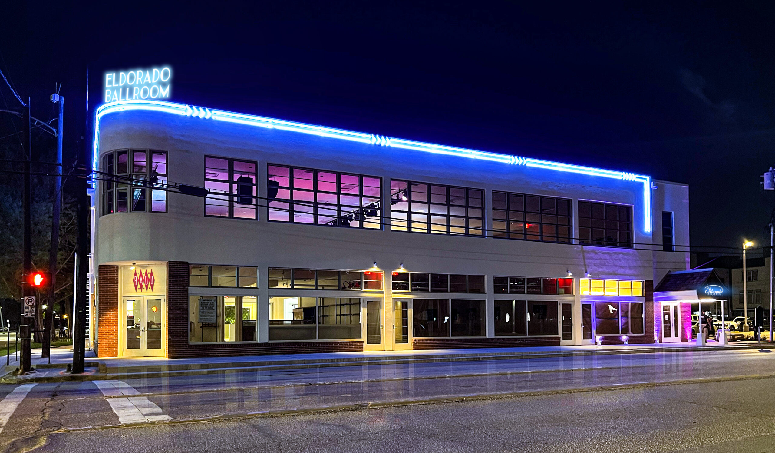 Nighttime image of a 2 story white building with a row of ribbon windows across the 2nd floor that wrap around the corner of the building. Blue neon lights accent the roof line and match the neon on a rectangular sign on the building's corner that says Eldorado Ballroom. The doorways on both ends of the building are lit, one with neon lights under a canopy.