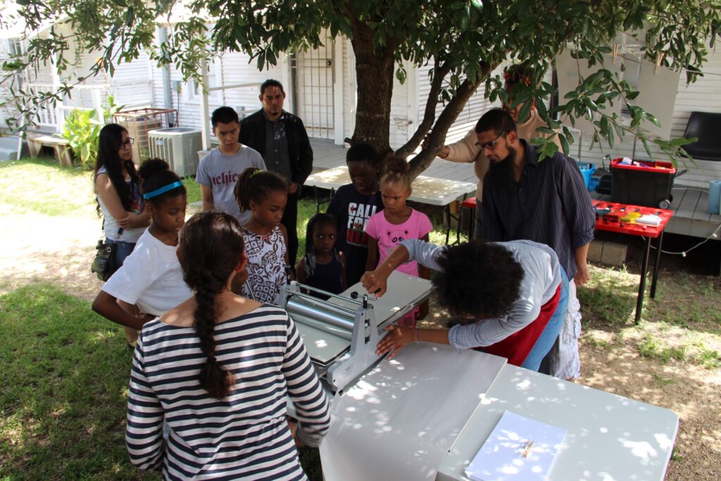 Children gathered around an instructor in a print-making workshop held outside in the courtyard under a tree.
